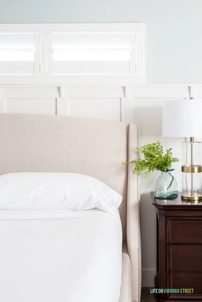 White bedding on neutral bed with a wood side table and clear glass vase with greenery in it.