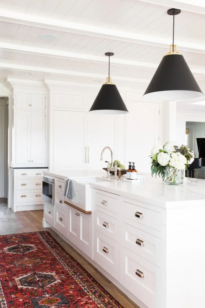 A mostly white kitchen with a red ornate rug and black lights hanging over the island.