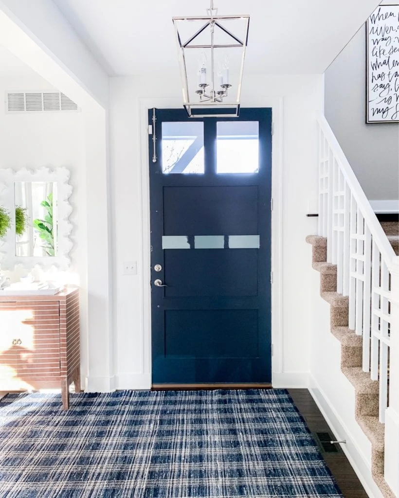 An interior front door painted Benjamin Moore Hale Navy with three blue gray paint swatches painted on the door. Also shows a navy blue plaid rug and silver pendant chandelier light next to a staircase.