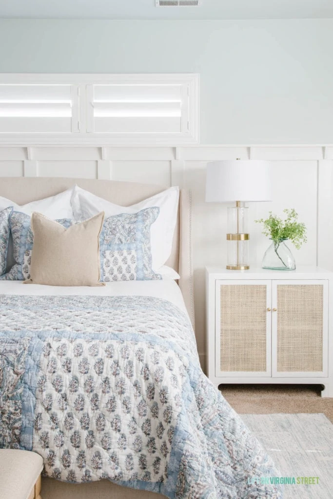 A bedroom with Sherwin Williams Sea Salt walls, a white and cane nightstand, a gold and glass lamp, blue paisley bedding and touches of greenery.