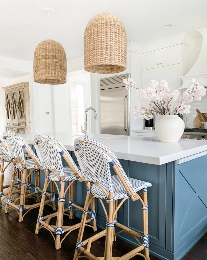 A pretty kitchen with a blue island, white cabinets, navy blue and white bistro counter stools, woven basket pendant lights, and faux pink cherry blossom stems!