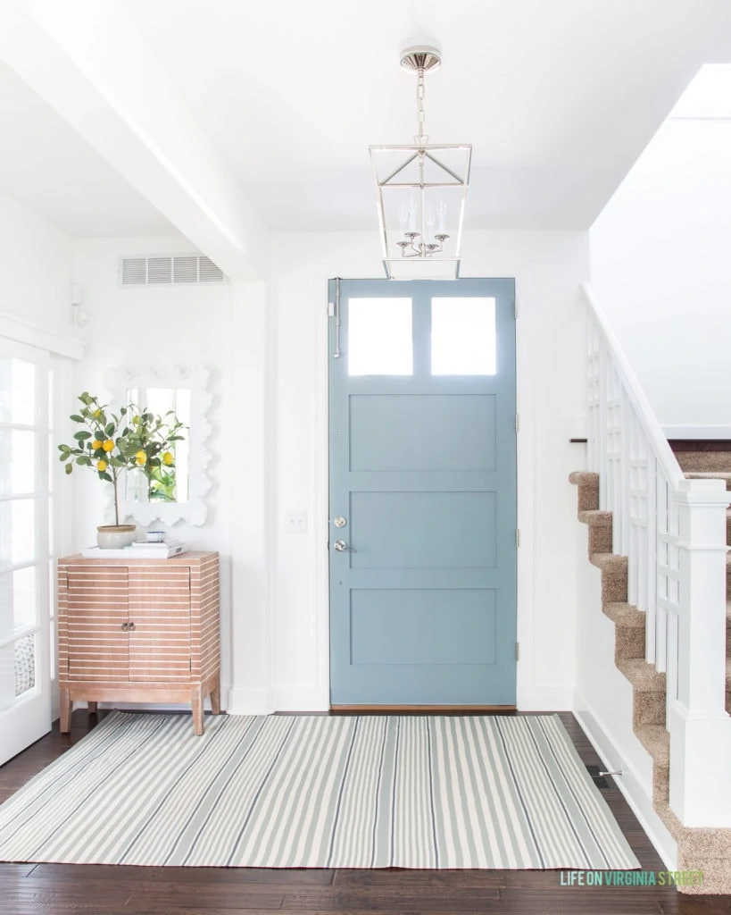 A beautiful entryway with a coastal vibe. Includes white walls, a blue gray interior painted door, striped rug, striped wood cabinet, a faux lemon tree topiary and a chrome lantern pendant light.