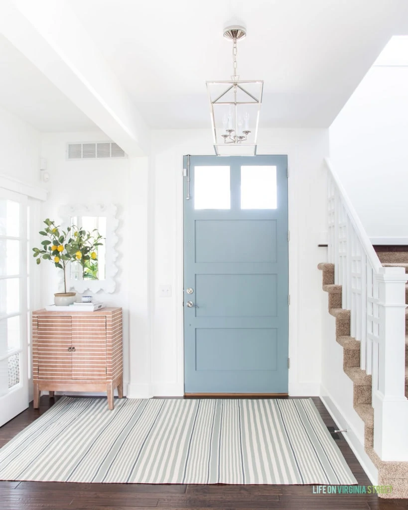 An entryway with Benjamin Moore Simply White Walls, BM Water's Edge blue door color, a wood striped cabinet, lemon tree topiary and a striped rug.