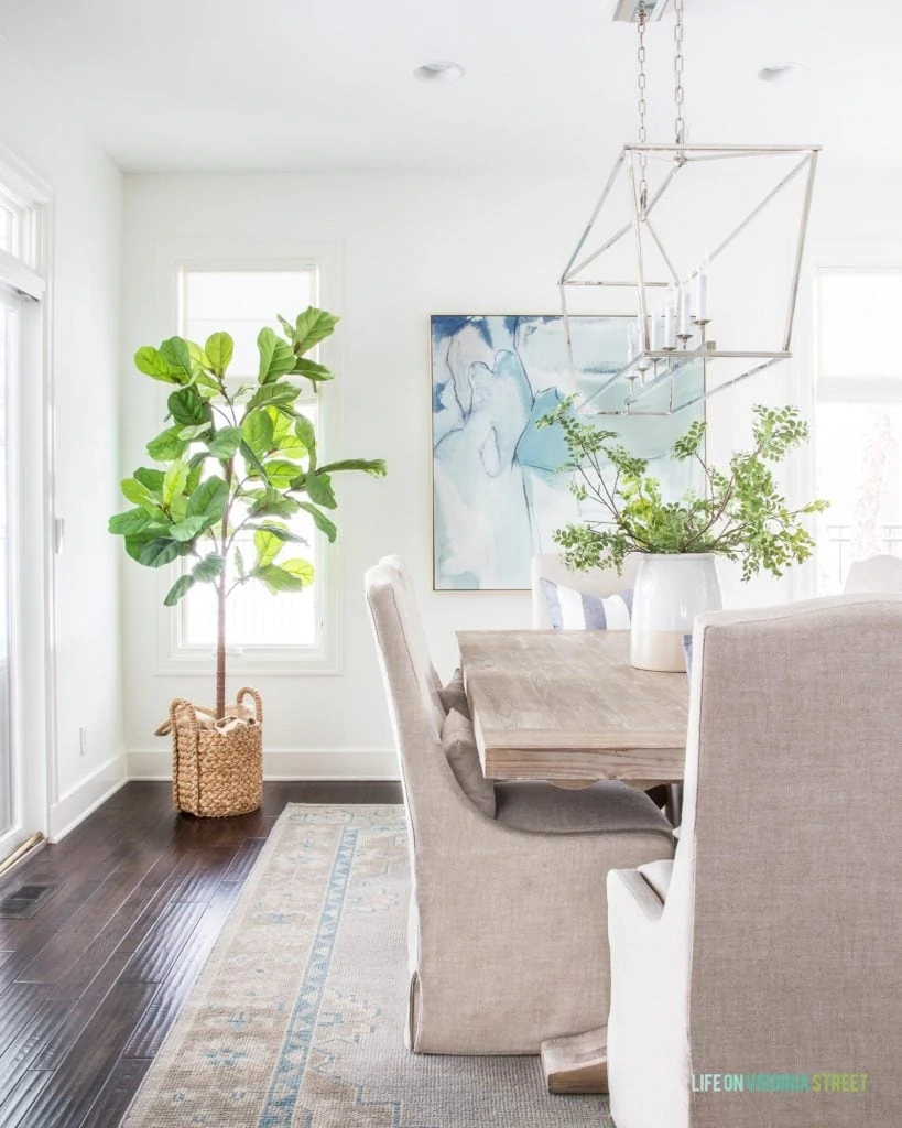 A coastal dining room with linen chairs, reclaimed wood dining table, blue abstract art, a chrome Darlana linear chandelier, hand knotted rug, faux fiddle leaf fig tree and white walls. This post shares how to get a similar look for less in your own home!