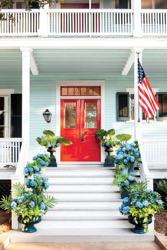 An aqua home with a bright red front door and large porch. The steps leading to the home are filled with potted blue hydrangeas.