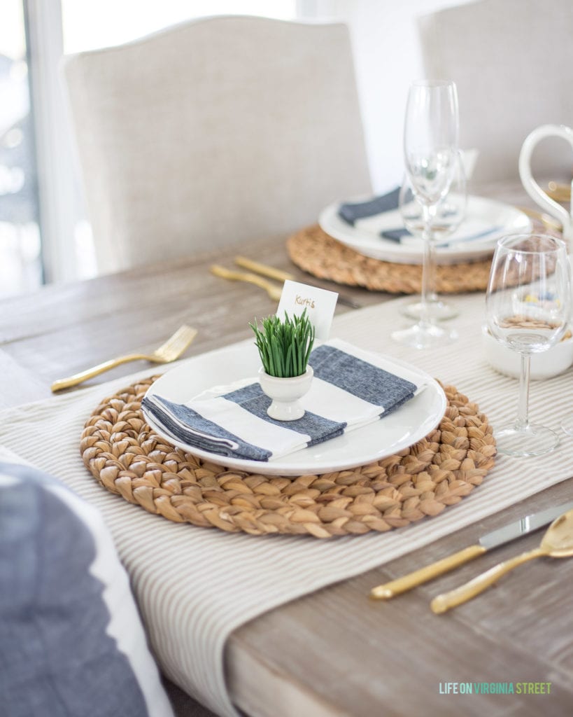 An Easter place setting with water hyacinth chargers, striped table runner, wide navy blue striped napkins, a grass place card holder and linen dining chairs!