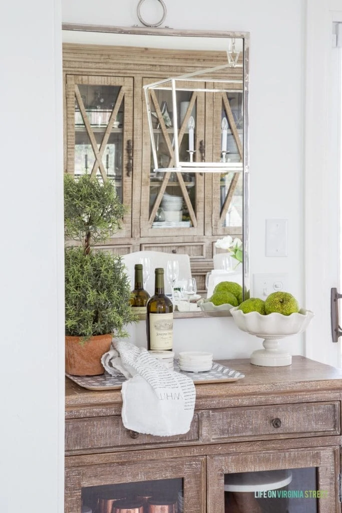 A wood buffet table with a silver mirror, faux topiary, and view of a spring dining room.