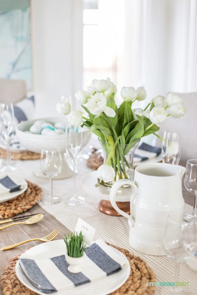An Easter tablescape with white ceramic serving pieces, a faux white tulip flower arrangement, sisal chargers and striped napkins.