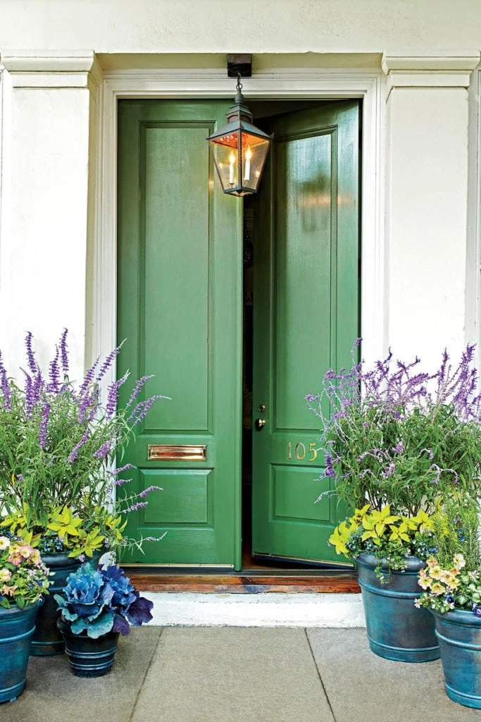 A beautiful bold green front door that is slightly ajar paired with planters filled with purple flowers and green foliage.