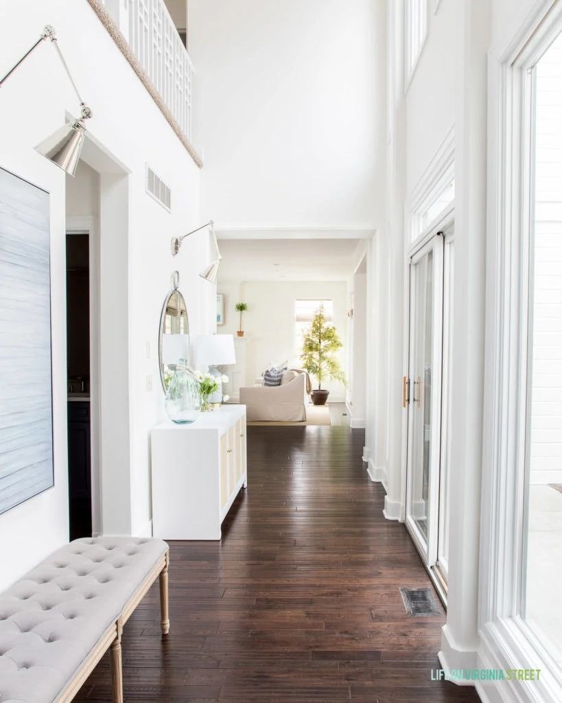 A bright entryway hallway with floor to ceiling windows, a gray tufted bench, white cane cabinet, swing arm sconces, a round mirror and tulips for spring!