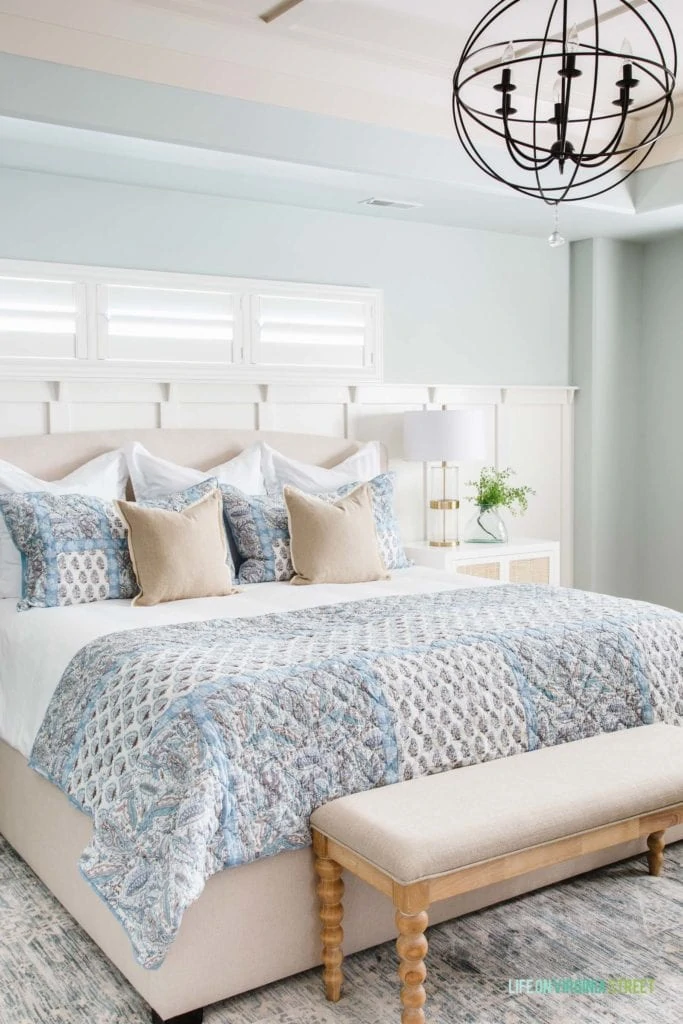 A large neutral bed with a blue and white quilt at the end of the bed and blue, white and neutral pillows.