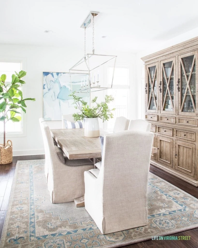 A spring dining room with a vintage style rug, linen dining chairs, a reclaimed wood dining table, linear chandelier and blue and green abstract art.