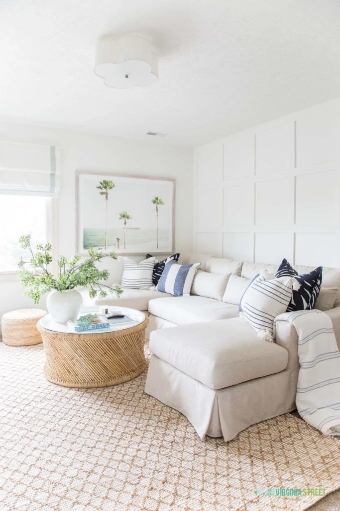 A light and bright den with jute rug, linen sectional, board and batten walls, a bamboo and enamel coffee table, palm tree art, ribbon trimmed roman shades and blue and white striped pillows.