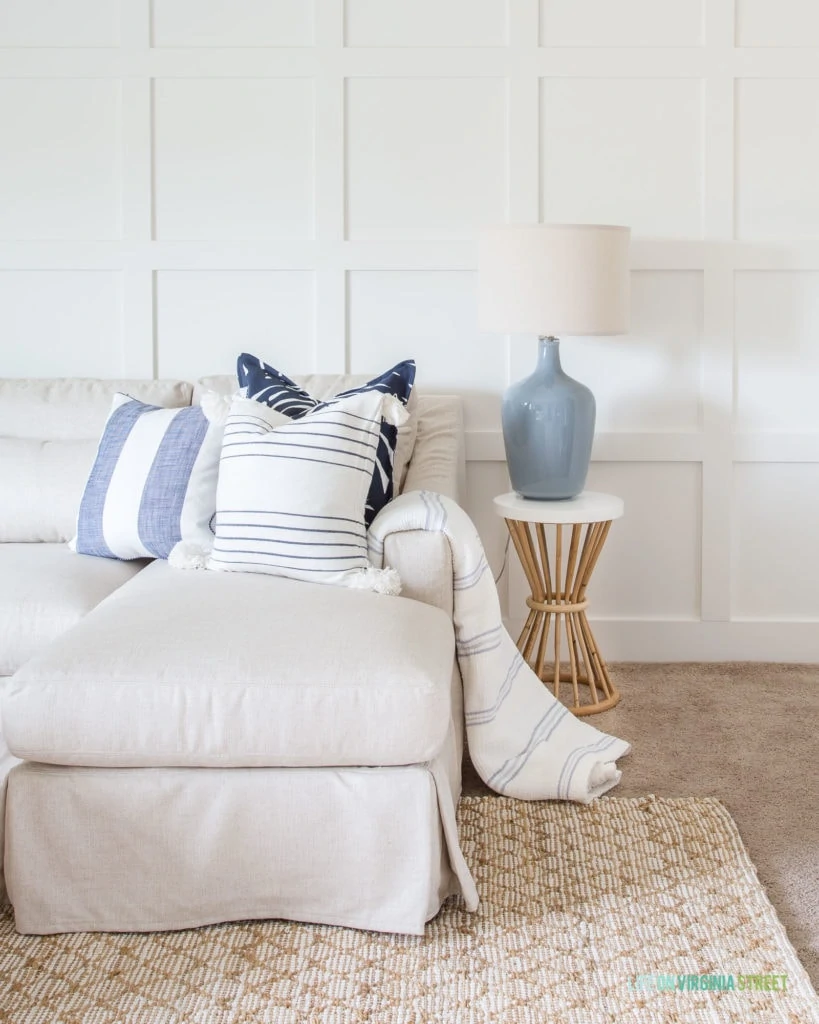 A white board and batten grid wall with a linen sectional, blue gray lamp, bamboo and white resin side table, and blue and white striped pillows.