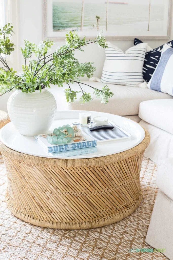 A coastal bamboo coffee table with white enamel tray top that is durable and great for indoor or outdoor use! Decorated with a vase of greenery, a blue and white linen tray for TV remotes, and various beach inspired books and glass beads.