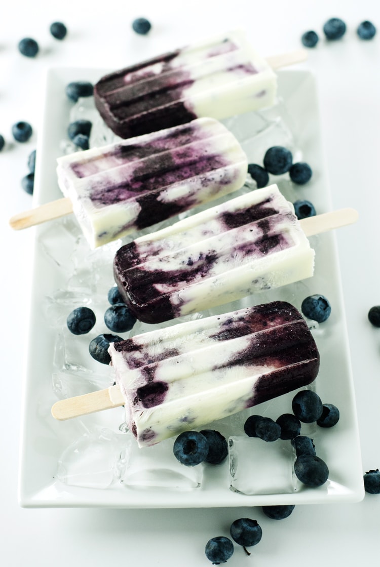 Blueberries and Cream Popsicles on the plate with blueberries scattered around them.