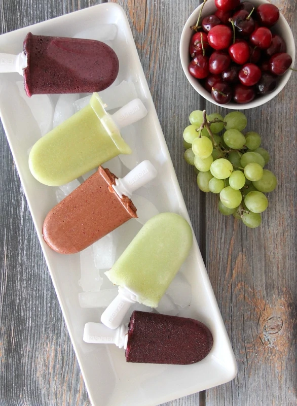 Easy Fruit Popsicles lying on a white plate and a bowl full of cherries and green grapes beside it.