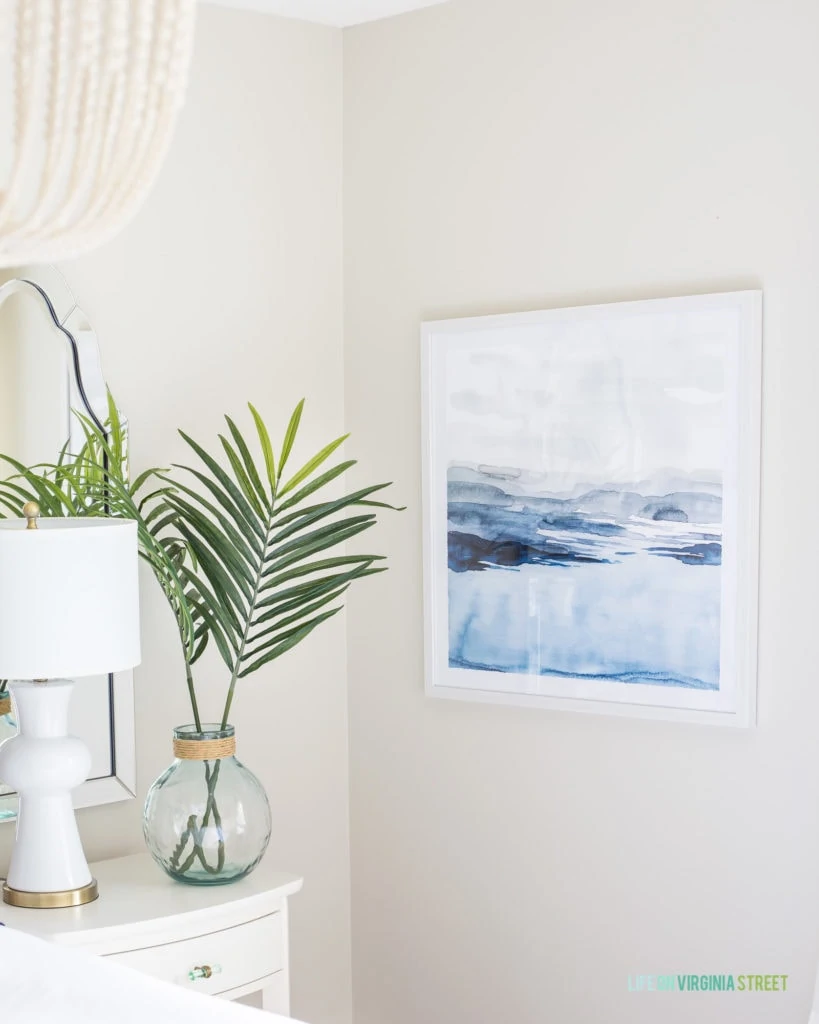 A clear vase on the side table in the bedroom with palm fronds in it and a beachy picture on the wall.