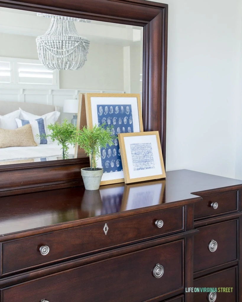 A dark wood dresser with framed pictures on it. A small topiary sits on top of the dresser and a chandelier is refelected in the mirror.