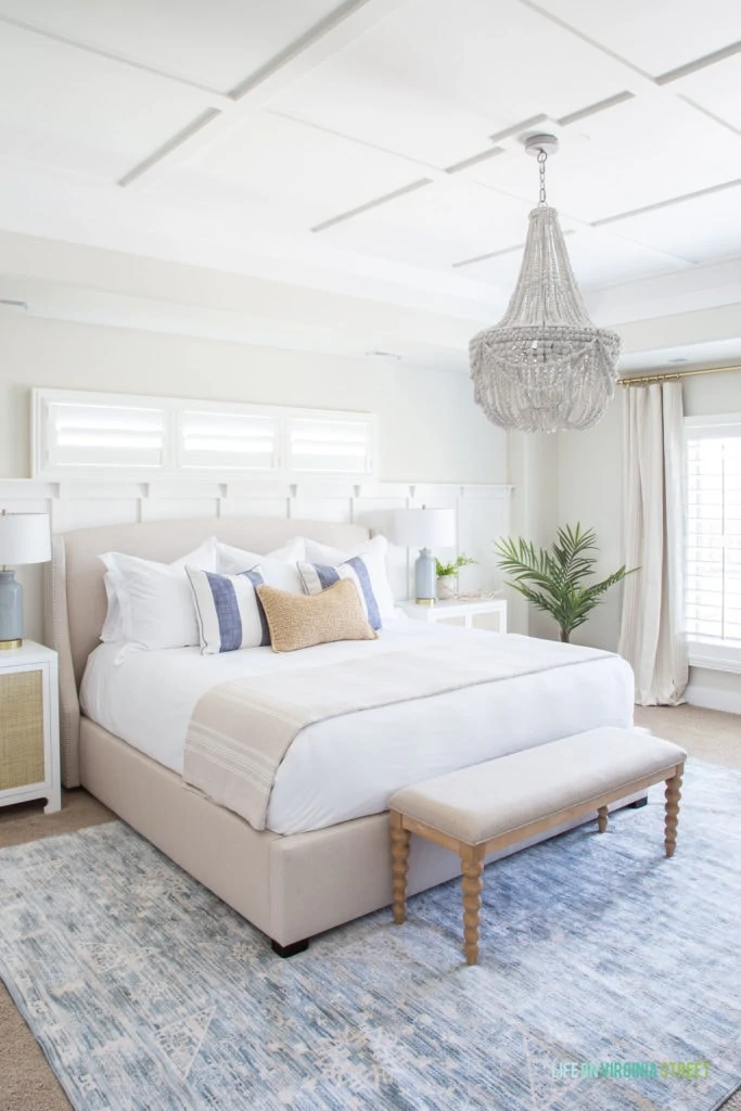 A large bedroom with white bedding, a small light blue rug and a beaded chandelier.