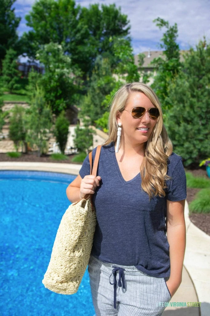 A woman standing beside the pool in aviator sunglasses, holding a beach bag.