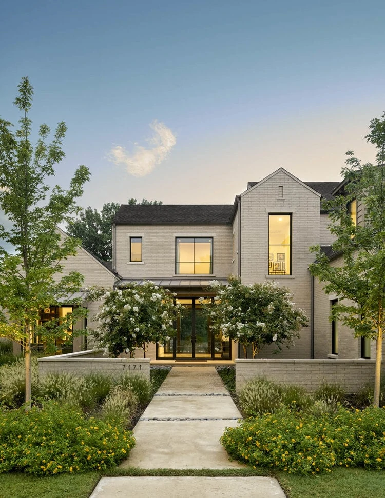 A light beige house with a cement walkway, trees on both sides leading to sliding doors of the house.