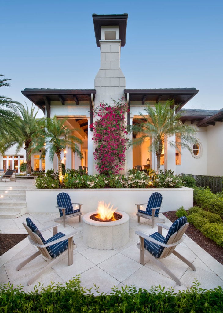 A beautiful Mediterranean-inspired outdoor patio with a white outdoor fireplace, firepit, concrete pavers and palm trees and bougainvillea.
