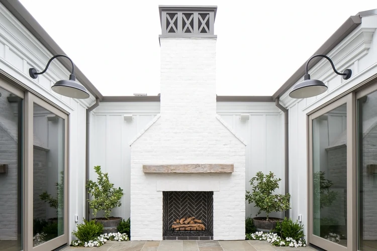 A white house with a brick outdoor fireplace, gray trim, large gray sliding doors, and barn lights in this area full of outdoor courtyard ideas.