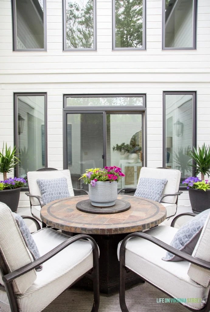A brown fire pit table in a courtyard with a white house, black window trim, and coastal inspired accessories.
