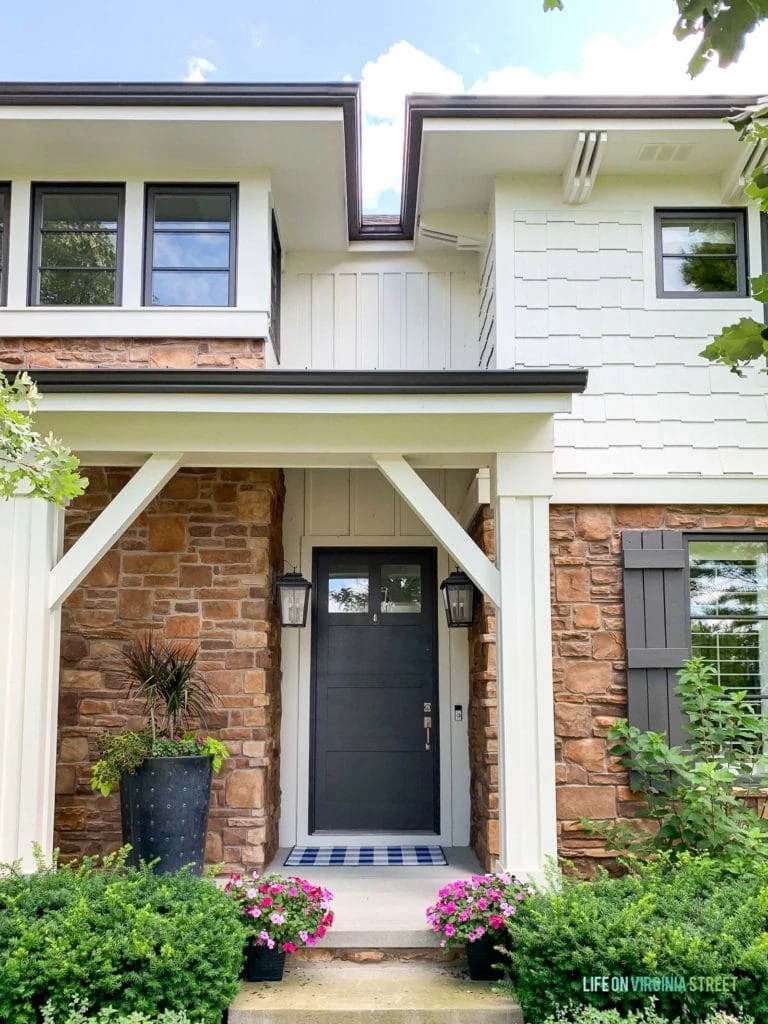 A front porch on a white house with brown stone and bronze window trim. Includes outdoor lantern light fixtures, a tall planter with flowers and a blue and white buffalo check rug.