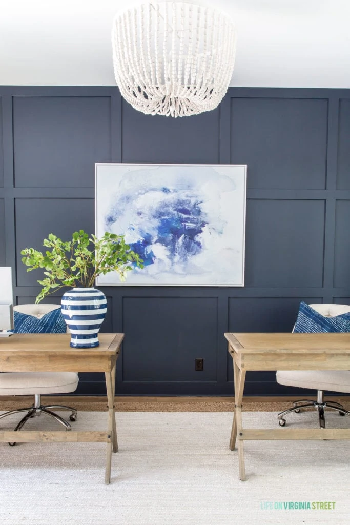 A home office with a navy blue board and batten accent wall, white and jute rug, wood desks, linen office chairs, white wood bead chandelier, blue and white abstract art, and a blue and white striped vase.