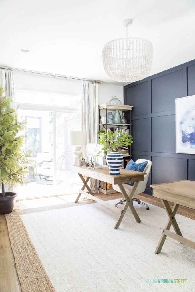 A light and bright home office with navy blue and white walls, jute rug, a pair of driftwood colored desks, white bead chandelier, wood and metal bookcases, striped drapes and white oak hardwood floors.