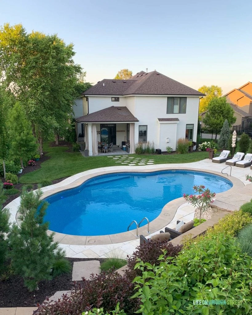 A white house with brown roof from the backyard view with an oasis shaped swimming pool.