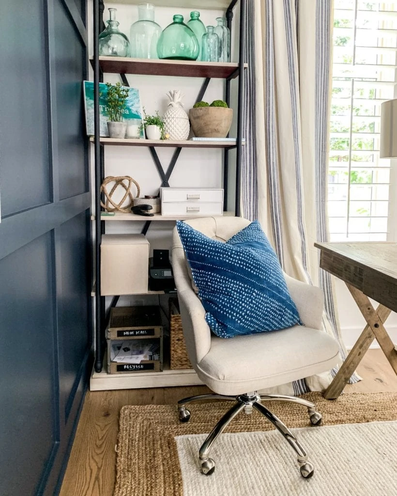 A home office space with navy blue wall, wood and metal bookcase, linen desk chair, jute rug, blue pillow and reclaimed wood desk.