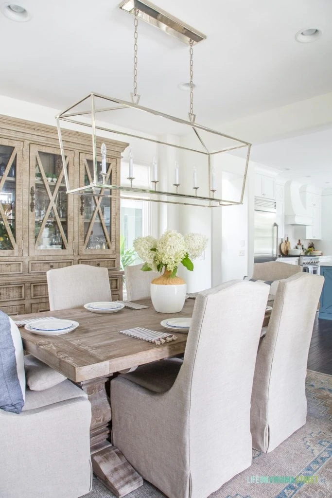 A neutral dining room with linen chairs, reclaimed wood table, wood hutch, silver Darlana linear chandelier, and blue and white accents. I love the limelight hydrangeas as the centerpiece on this tablescape!
