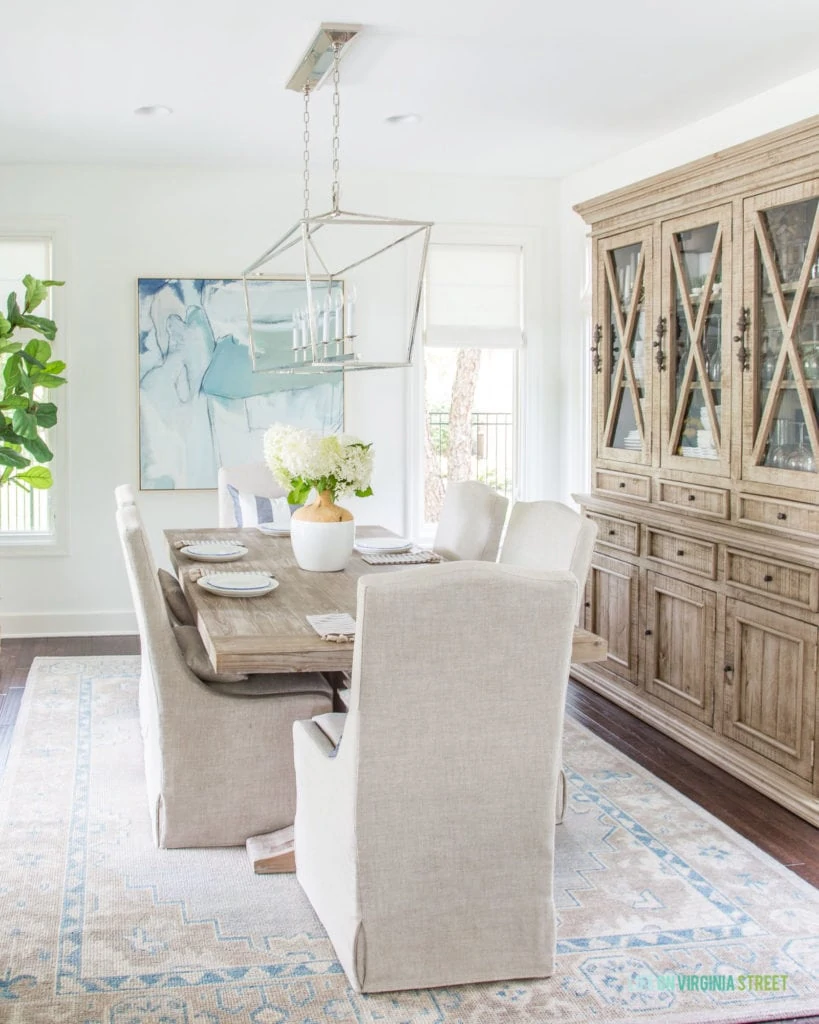 A coastal inspired dining room with a simple summer tablescape. I love the limelight hydrangeas, blue and white scalloped plates, striped tassel napkins, and blue and neutral accents in this space. Includes linen dining chairs, reclaimed wood dining table, blue abstract art, and a faux fiddle leaf fig tree.