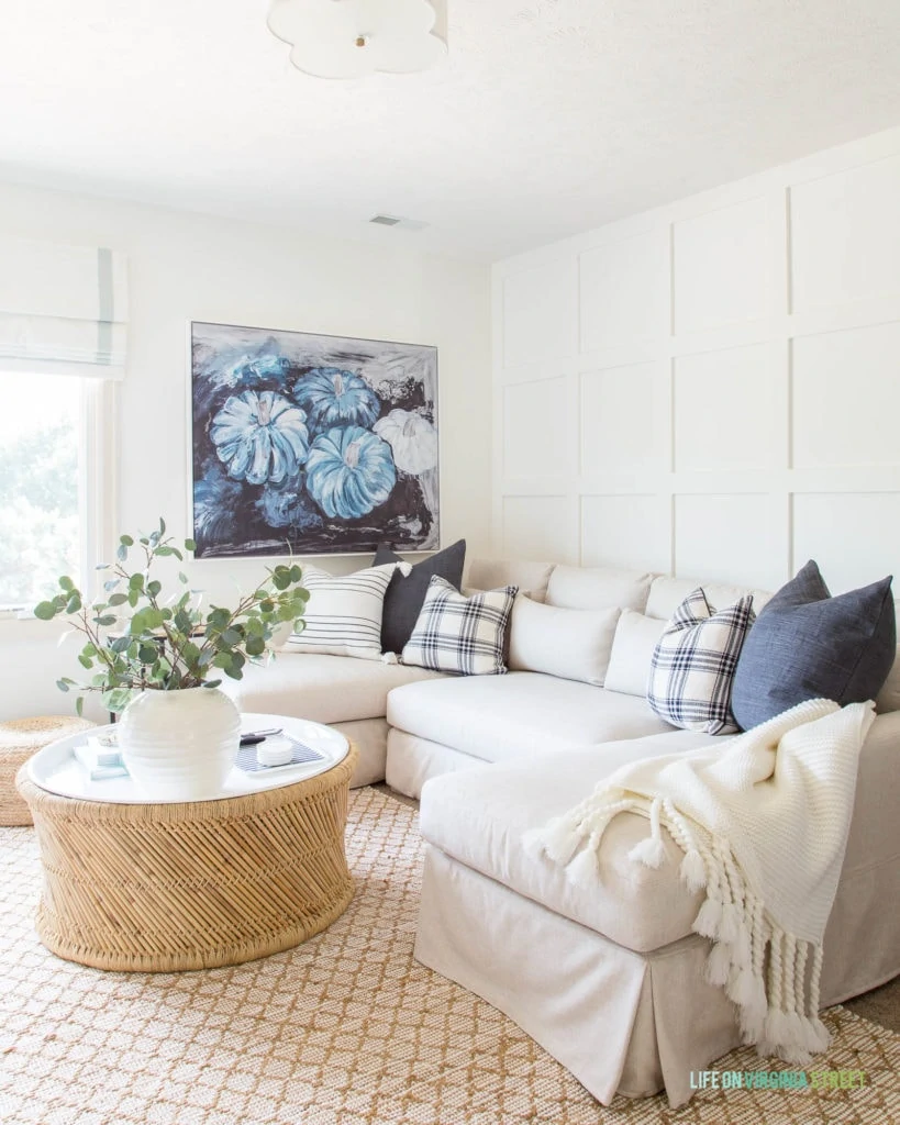 A fall living room with white board and batten grid wall, blue pumpkin abstract art, linen sectional sofa, jute rug, bamboo coffee table, and blue and white plaid pillows.