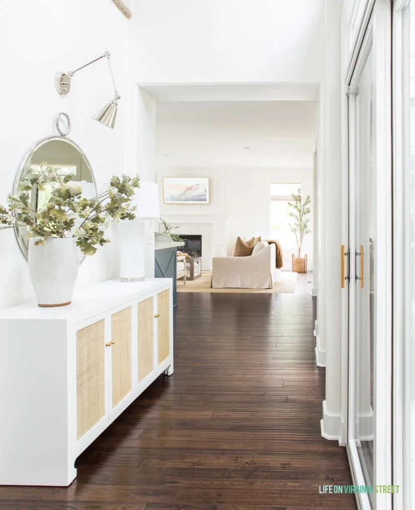 A tall white ceramic vase in an entryway with a white and cane console table and silver accents.