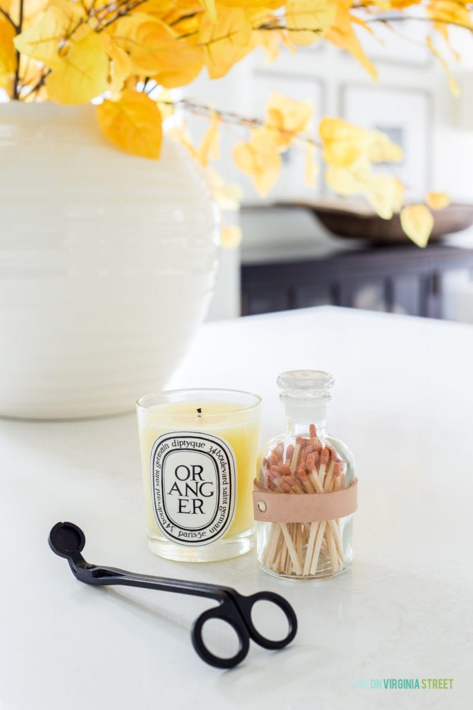 A countertop with a fall candle, wick trimmer, jar of matches, and a white vase filled with aspen leaves.