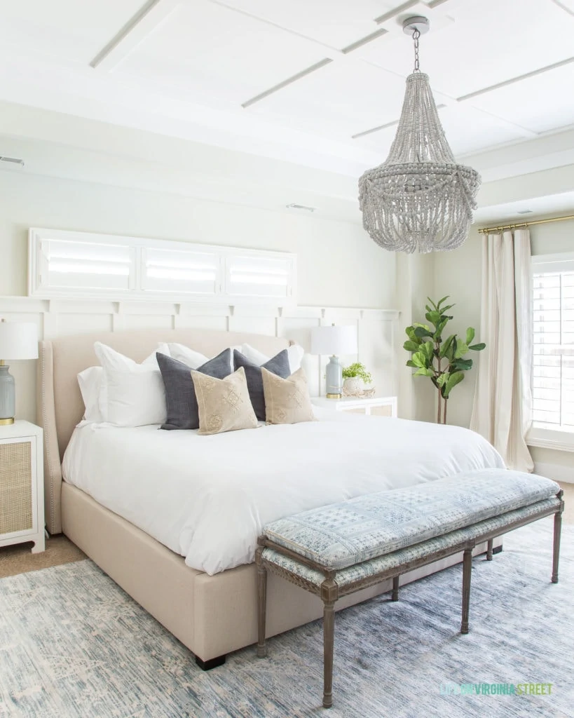 A coastal inspired master bedroom with white and cane nightstands, white wood bead chandelier, blue and white patterned bench with wood legs, white bedding, and blue and white accents.