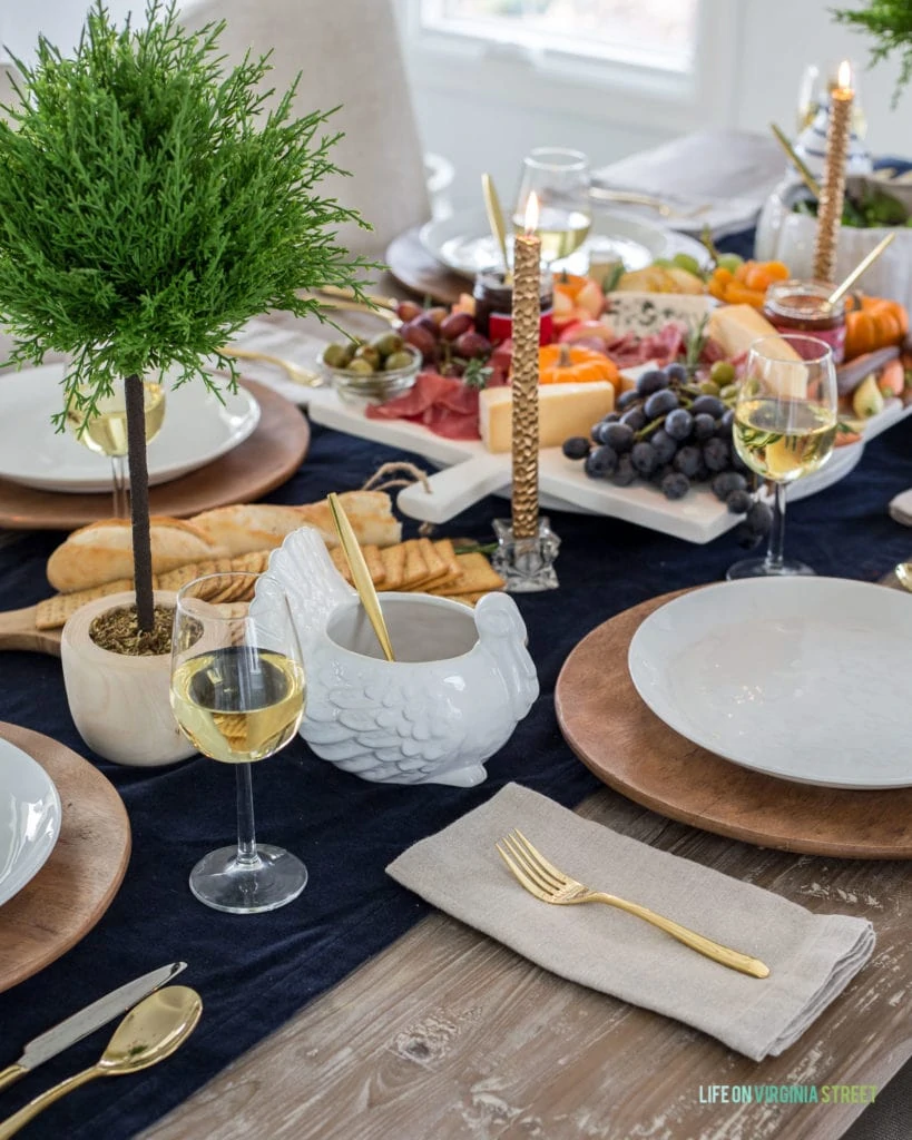 A simple and beautiful Thanksgiving table setting with a navy blue velvet table runner, white ceramic turkey gravy bowl, gold flatware, wood chargers, linen napkins and faux cypress topiaries. So pretty!