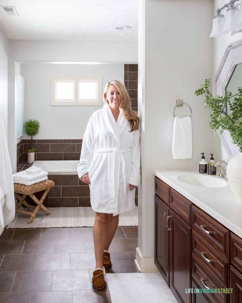 A woman in a white robe in the bathroom leaning against the wall.