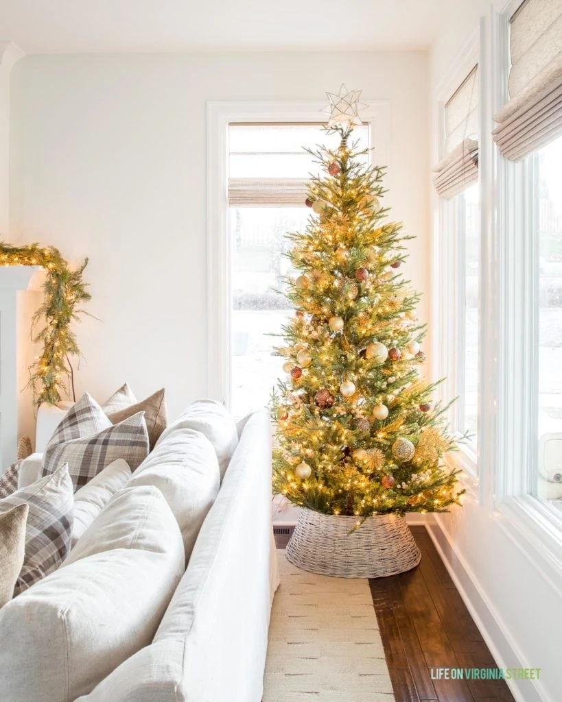 A beutiful neutral living room decorated for Christmas with a natural looking Christmas tree, whitewashed woven tree collar, a linen sofa, and neutral plaid pillows.