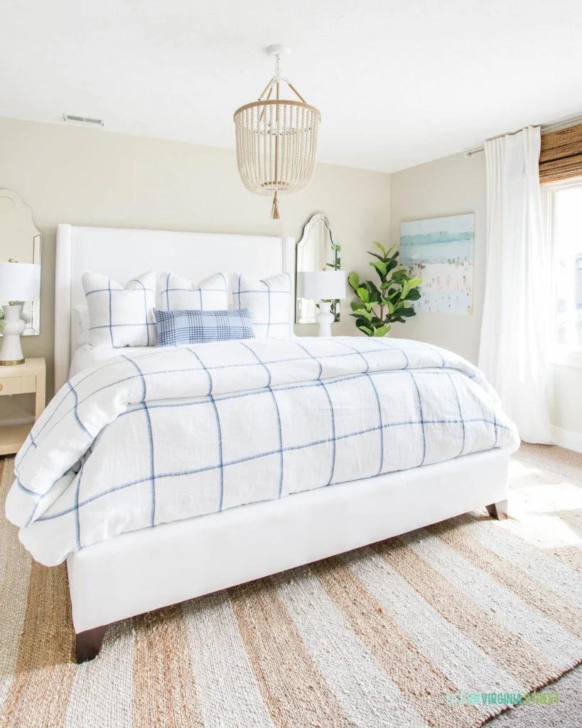 Abstract beach art hanging in a bedroom with a white upholstered bed, blue and white plaid bedding, a white bead chandelier, and neutral striped rug.