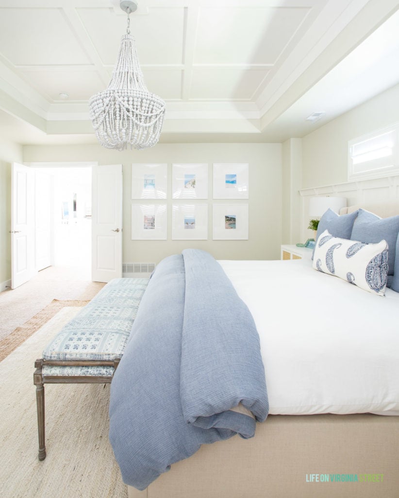 An oversized white photo gallery wall in a master bedroom with blue and white decor. Includes a white bead chandelier, blue bedding, and jute rug.