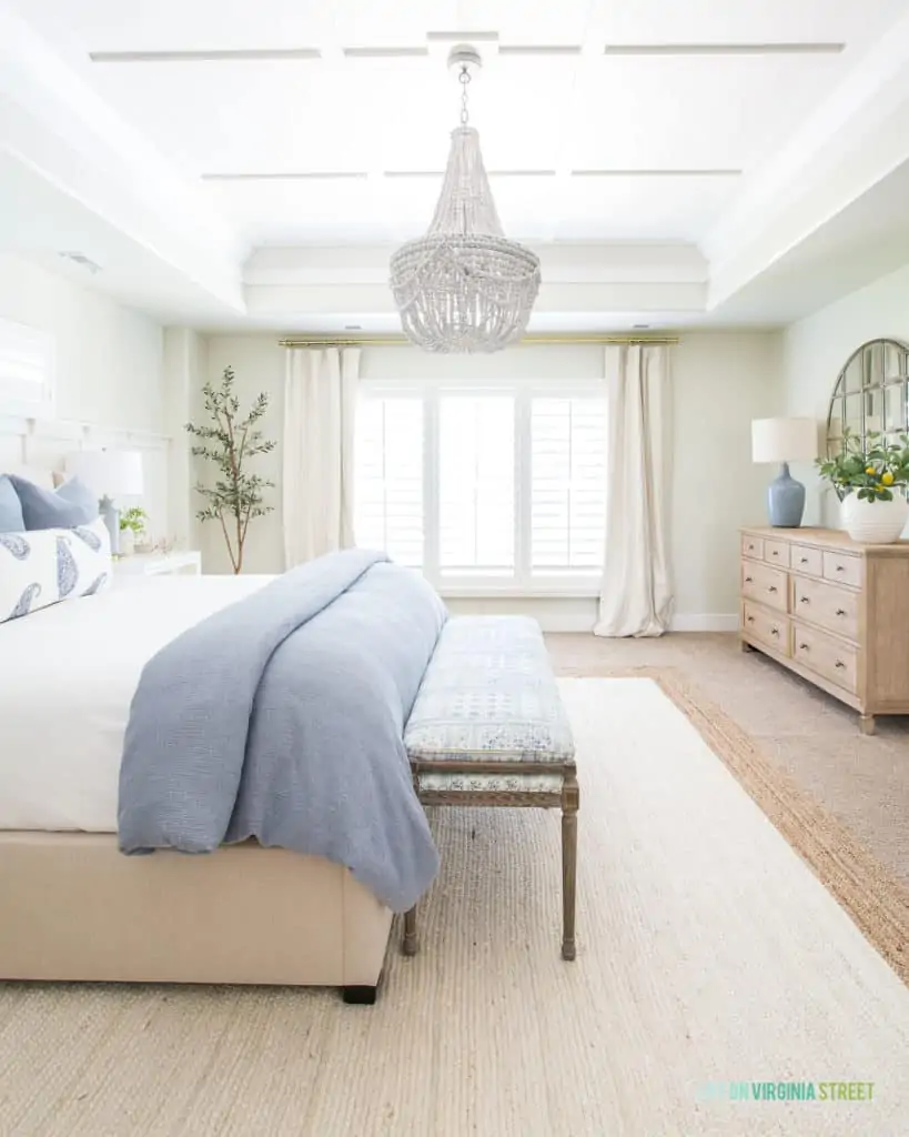 A serene bedroom with a white jute neutral area rug, blue bedding, a white wood bead chandelier, light wood dresser and plantation shutters on the windows.