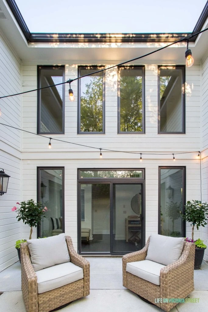 A courtyard patio with wicker swivel chairs, string lights, and a white house with black window trim.