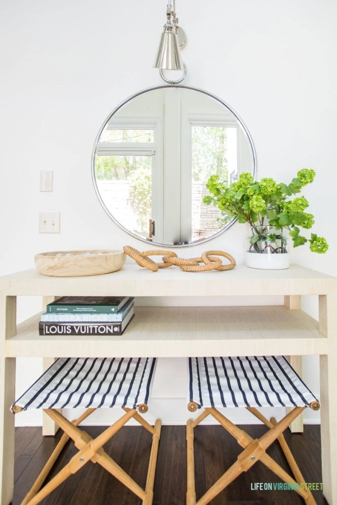 A light raffia console stable styled with a wood bowl, wicker chain, faux viburnum stems, and books. There is a round mirror over the table and striped x-benches below it.