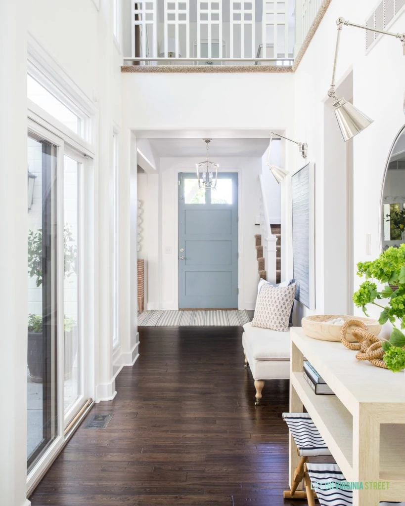 An entryway hallway with coastal decorating style. Includes a raffia console table, wicker chain decor, faux viburnum stems, a pillowtop bench, swing arm sconces, and a light blue gray interior door.