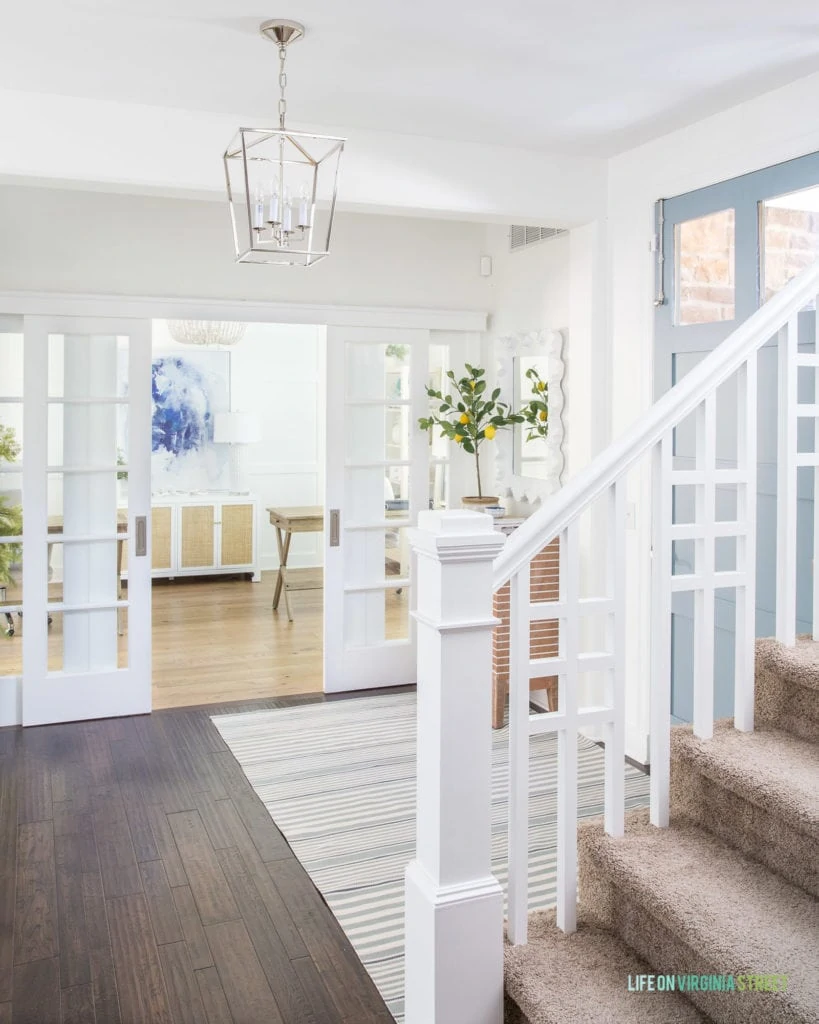 A coastal inspired entryway with a blue gray painted interior front door, a striped rug, white walls and a white board & batten grid wall in the background with a home office.